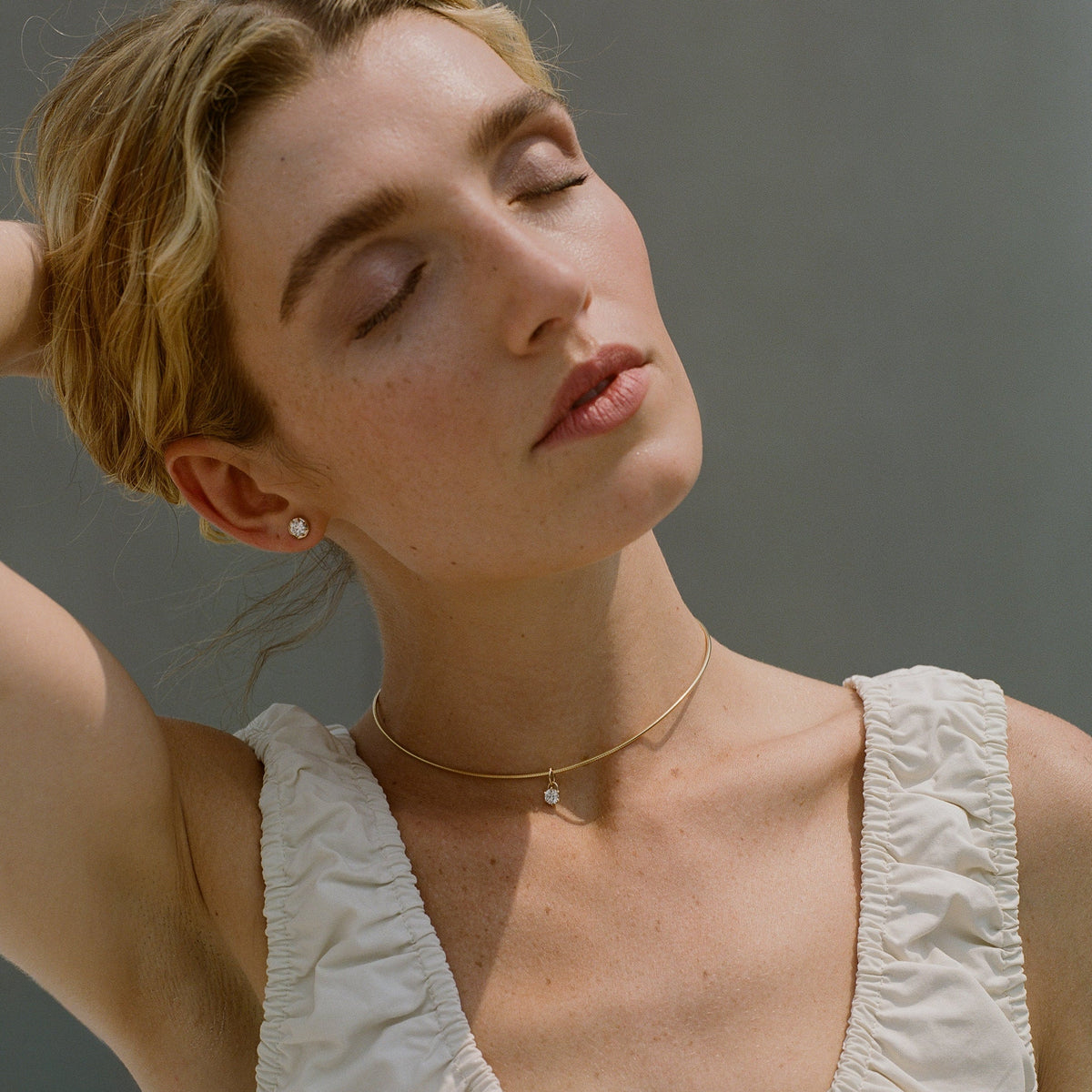 A close-up lifestyle shot of a woman with closed eyes wearing a delicate gold chain necklace with a tiny pearl-like bead pendant, paired with a white ruffled top against a neutral gray background.