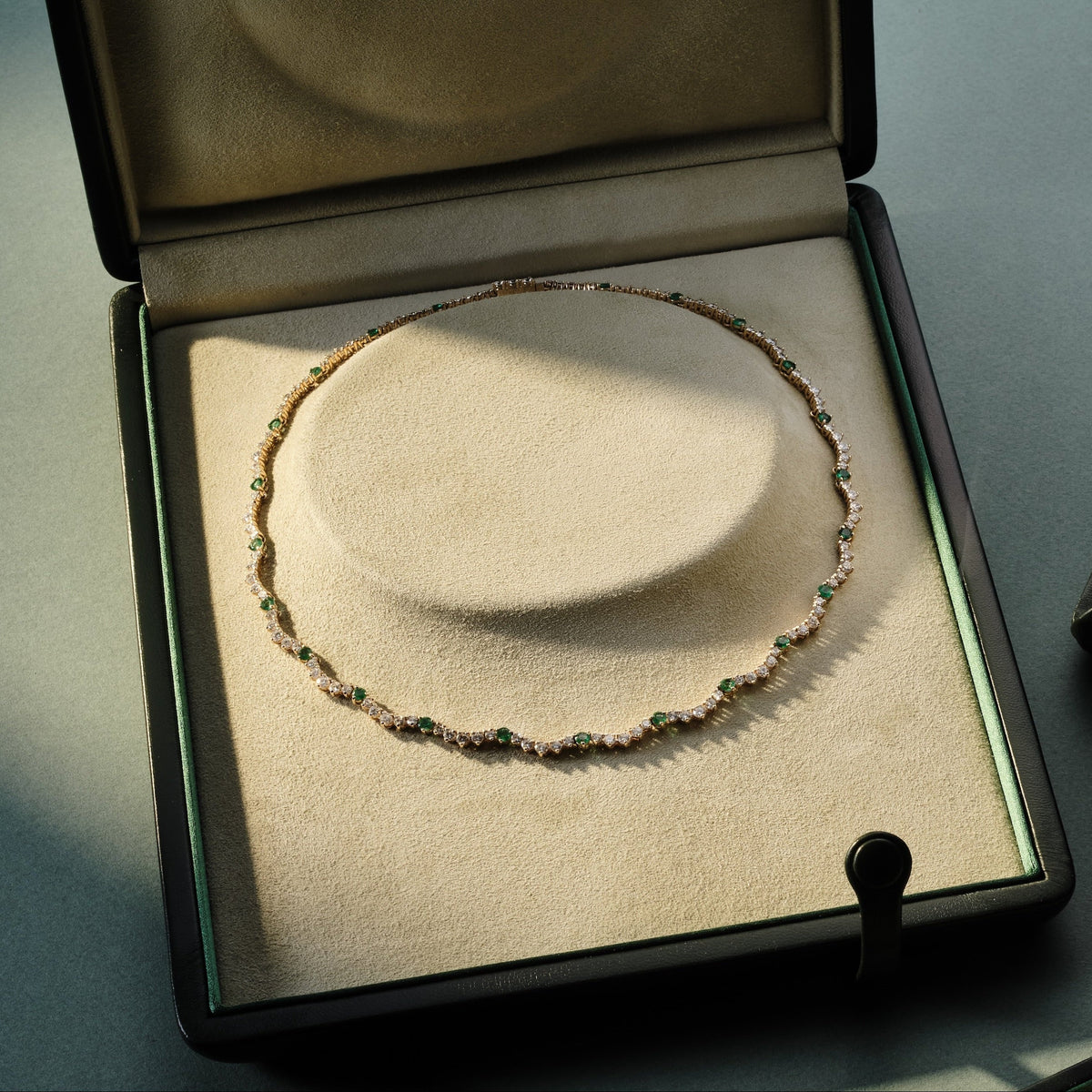 An open jewelry box displaying a delicate beaded necklace, alongside other open jewelry boxes containing a curved cuff bracelet and a gold chain accessory, arranged on a dark blue surface.