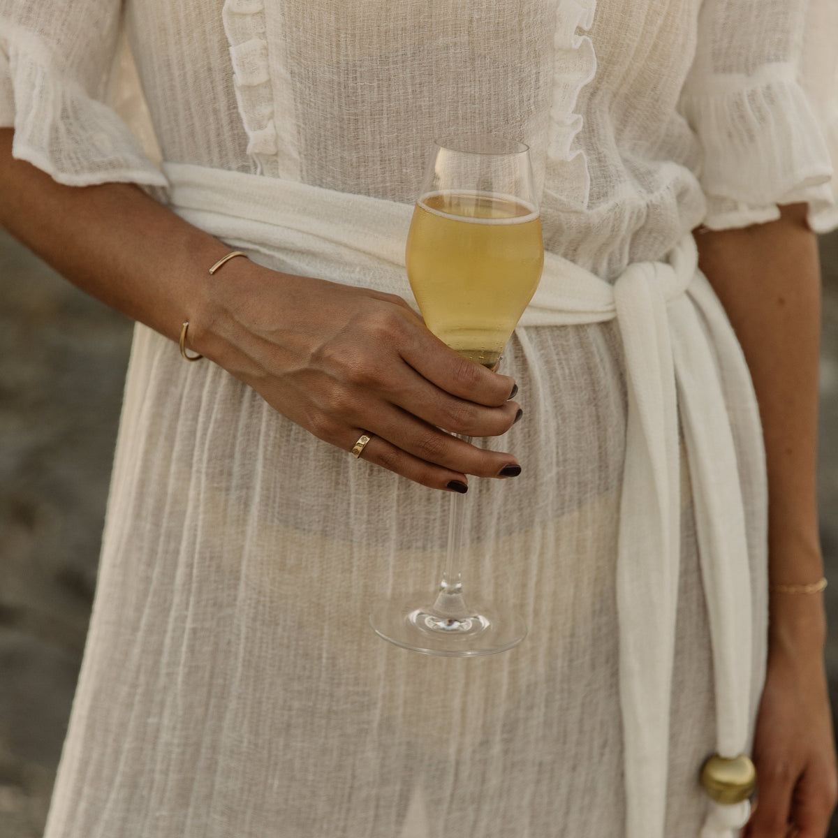 A close-up of a person wearing a flowing white or cream sheer maxi dress with ruffled trim and a tied waist sash. The person is holding a glass of pale yellow beverage, with gold jewelry visible, against a softly blurred outdoor background.
