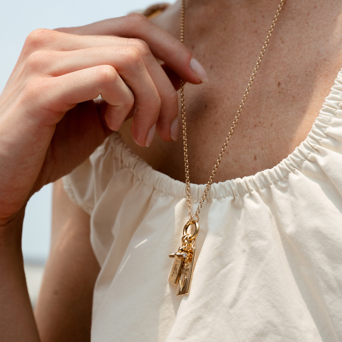 A close-up of a model wearing a delicate gold chain necklace with a small cross and rectangular pendant, styled with a white off-the-shoulder top.