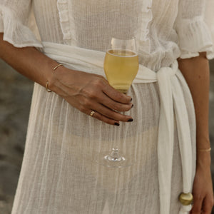 A close-up of a person wearing a flowing white or cream sheer maxi dress with ruffled trim and a tied waist sash. The person is holding a glass of pale yellow beverage, with gold jewelry visible, against a softly blurred outdoor background.
