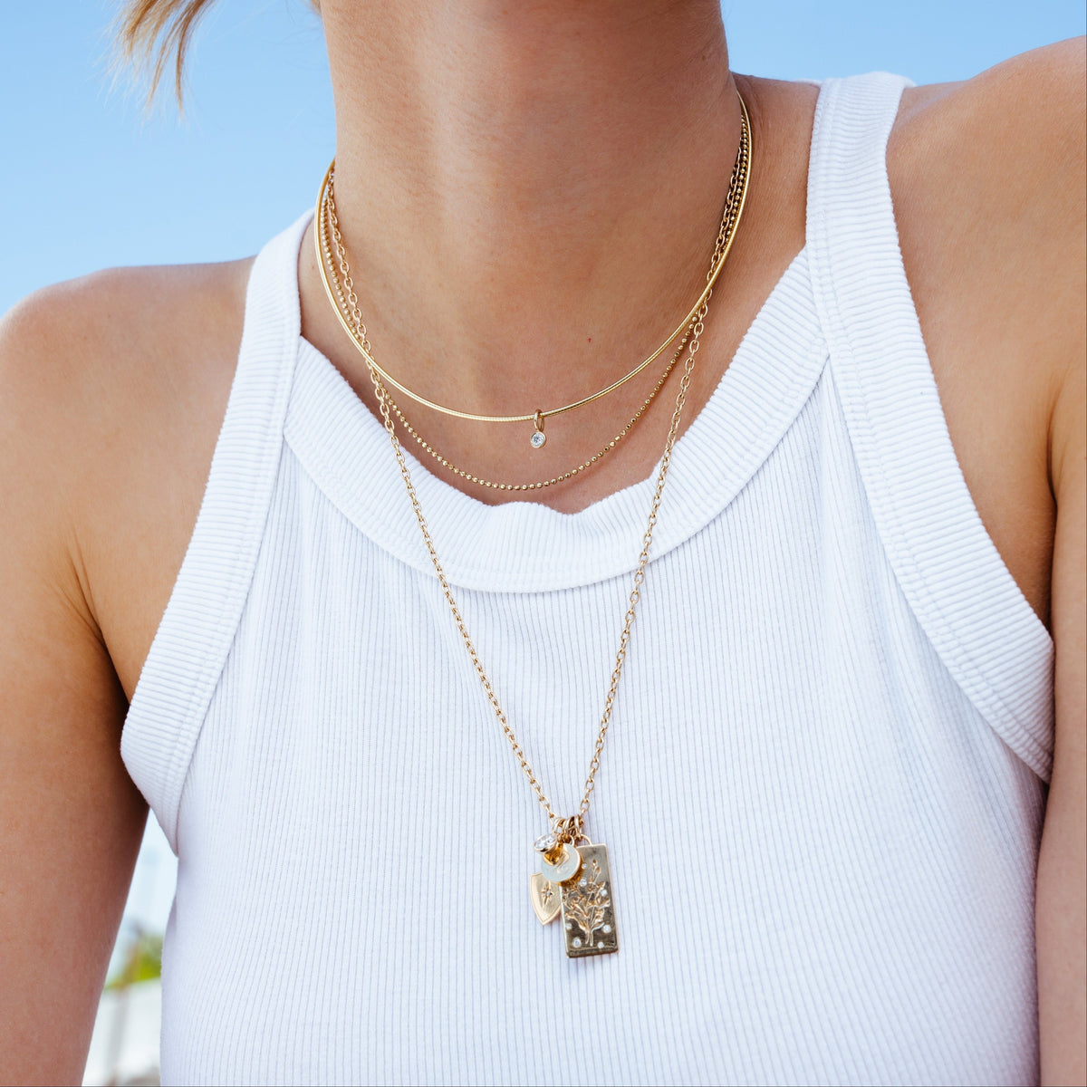 A close-up lifestyle shot of a woman wearing a layered gold necklace with a rectangular pendant featuring a small gold heart and a textured beige inlay, styled with a white ribbed tank top against a blue sky background.